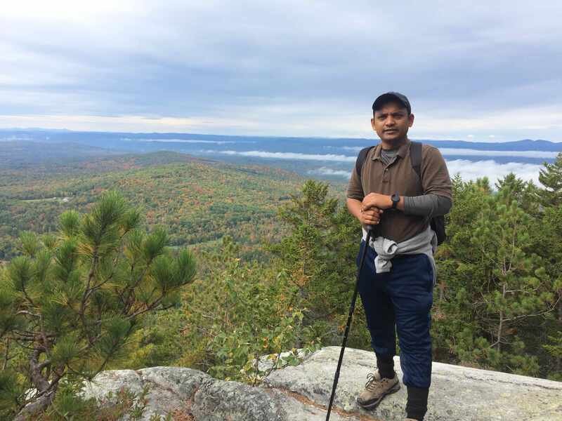 Arasu standing on a rocky overlook with a backpack and walking pole, forested hills behind him