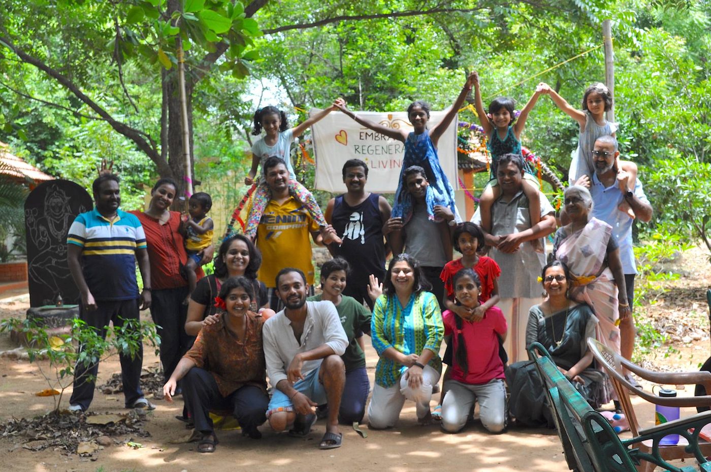 A large intergenerational group of adults and children standing and kneeling together outdoors under trees, holding a hand-painted banner reading Embracing Regenerative Living.
