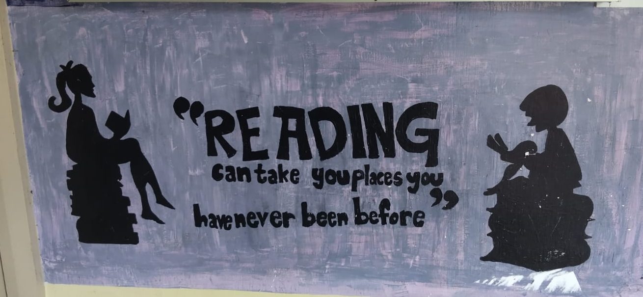 A hand-painted school mural showing two children reading, with the words: Reading can take you places you have never been before